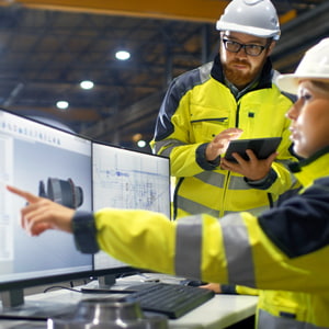 Inside a heavy industry factory, a female industrial engineer shows a colleague her design of a 3D turbine model on a dual-screen computer.